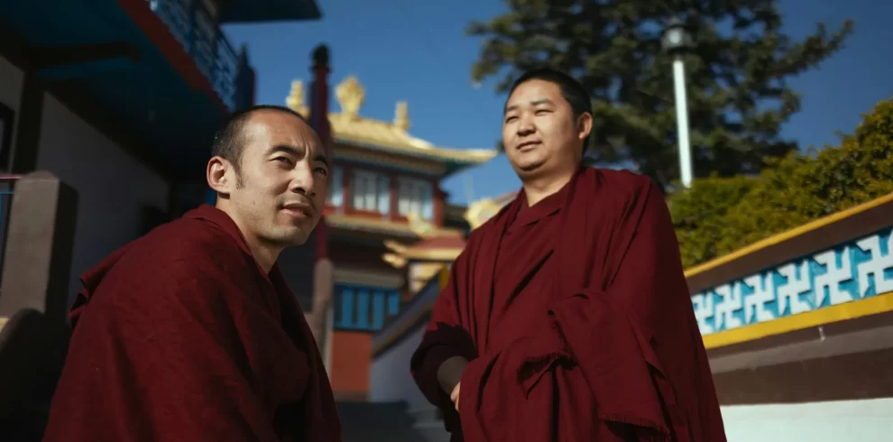 Two monks wearing maroon robes sitting outside a monastery.