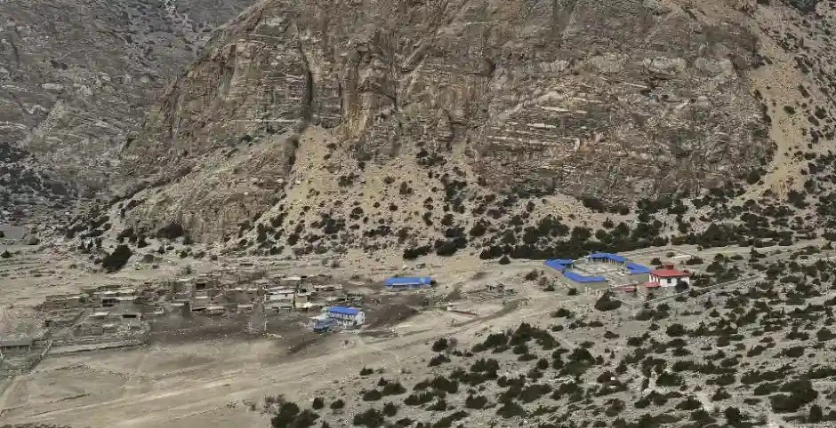 Kyang Village in Nar Phu Valley, a cluster of low buildings with some blue roofs situated at the base of a towering, steep, arid mountain cliff face.